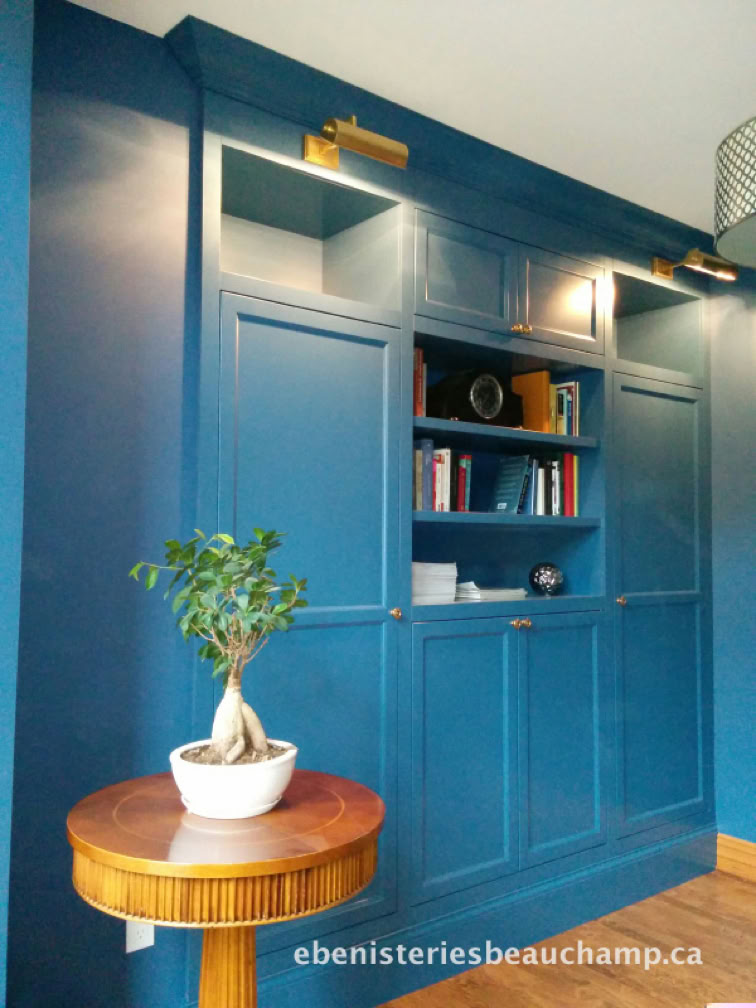 Blue built-in shelving unit with books and decorative items, illuminated by two lights, next to a small round table with a potted plant.