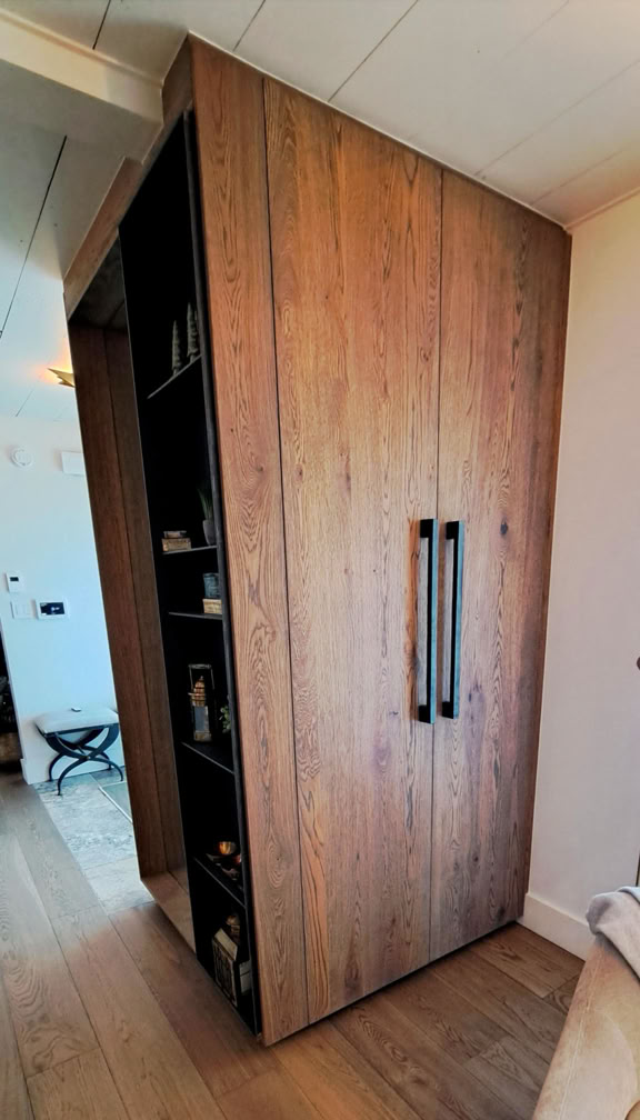 Wooden wardrobe with black handles, partially open to reveal shelves with various items inside, situated in a room with wooden flooring.