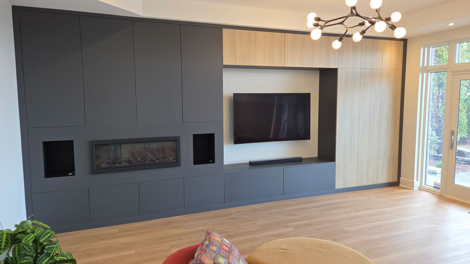 Modern living room with a large wall unit featuring a flat-screen TV and a built-in fireplace. The unit has dark and light wood panels. A contemporary chandelier hangs from the ceiling, and there are large windows on the right.